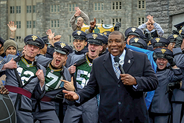 Curt Menefee, host of Fox NFL Sunday, speaks to the TV audience while cadets cheer behind him.